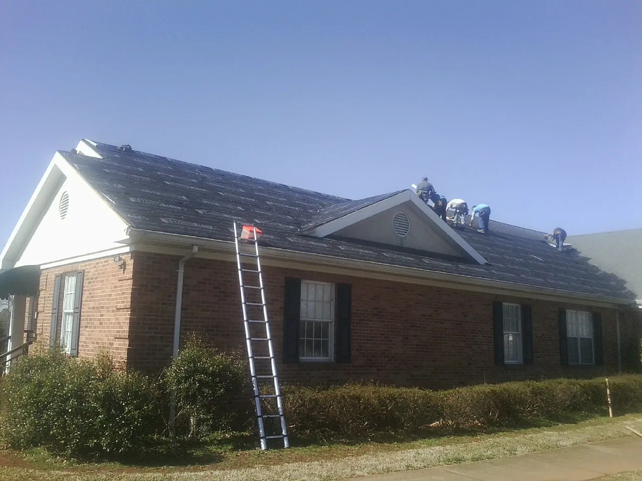 Crew tearing shingles off a brick ranch roof with a ladder set — Emergency Roof Repair work by our crew in Clemmons
