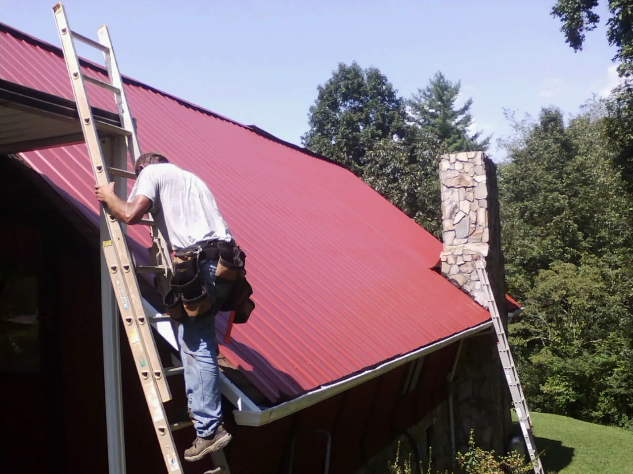 Roofer on a ladder beside a red metal roof and stone chimney — Soffit & Fascia Repair by Roofing Brothers in Clemmons